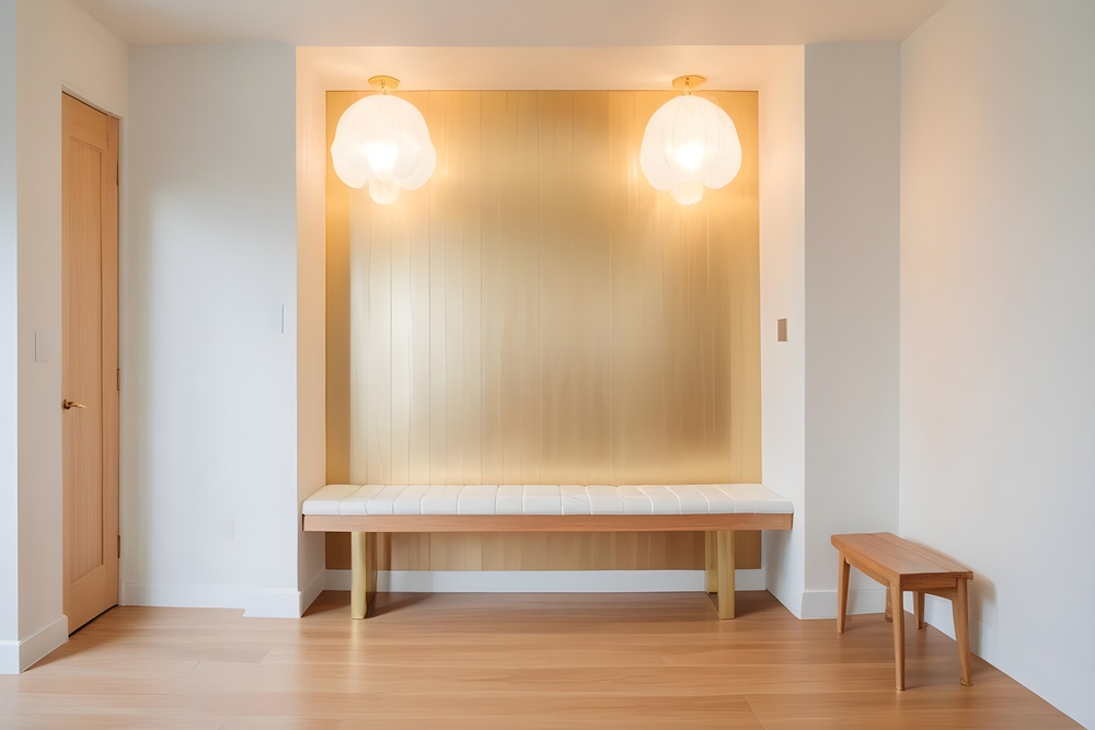 Recibidor decorado con una banqueta blanca y respaldo dorado, acompañado de iluminación cálida con lámparas de techo y un pequeño banco de madera, creando un espacio acogedor y elegante.